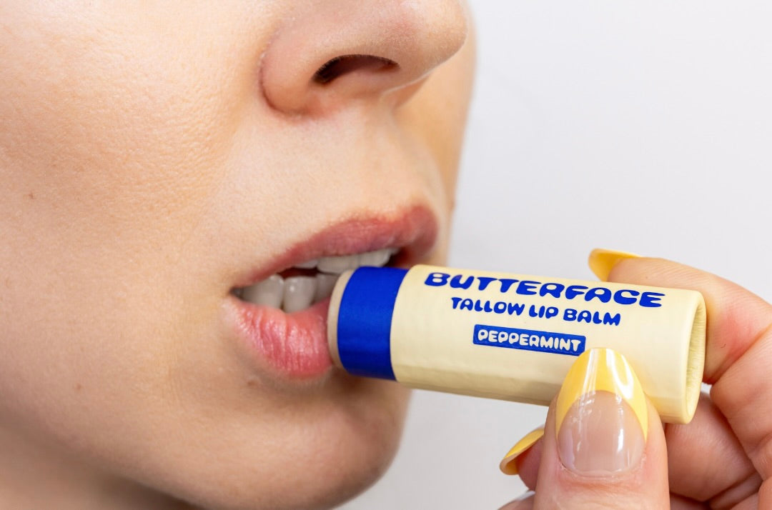 A close-up of a person applying Butterface Tallow Lip Balm in Peppermint from a cream-colored tube with blue accents, held by a hand with yellow French-tipped nails against a plain white background.