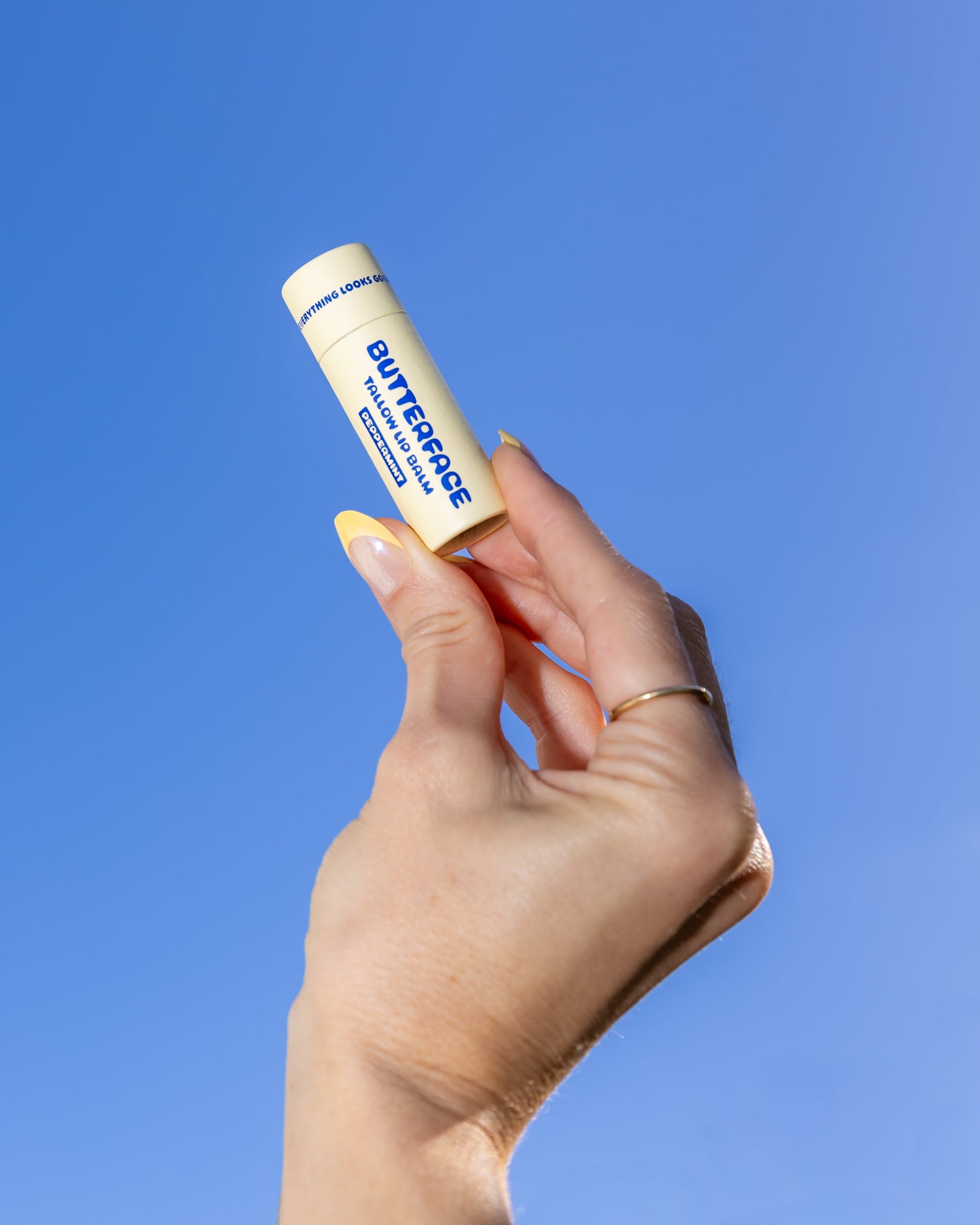 Hand holding a tallow butter lip balm named Butterface in a plastic-free tube against a blue sky