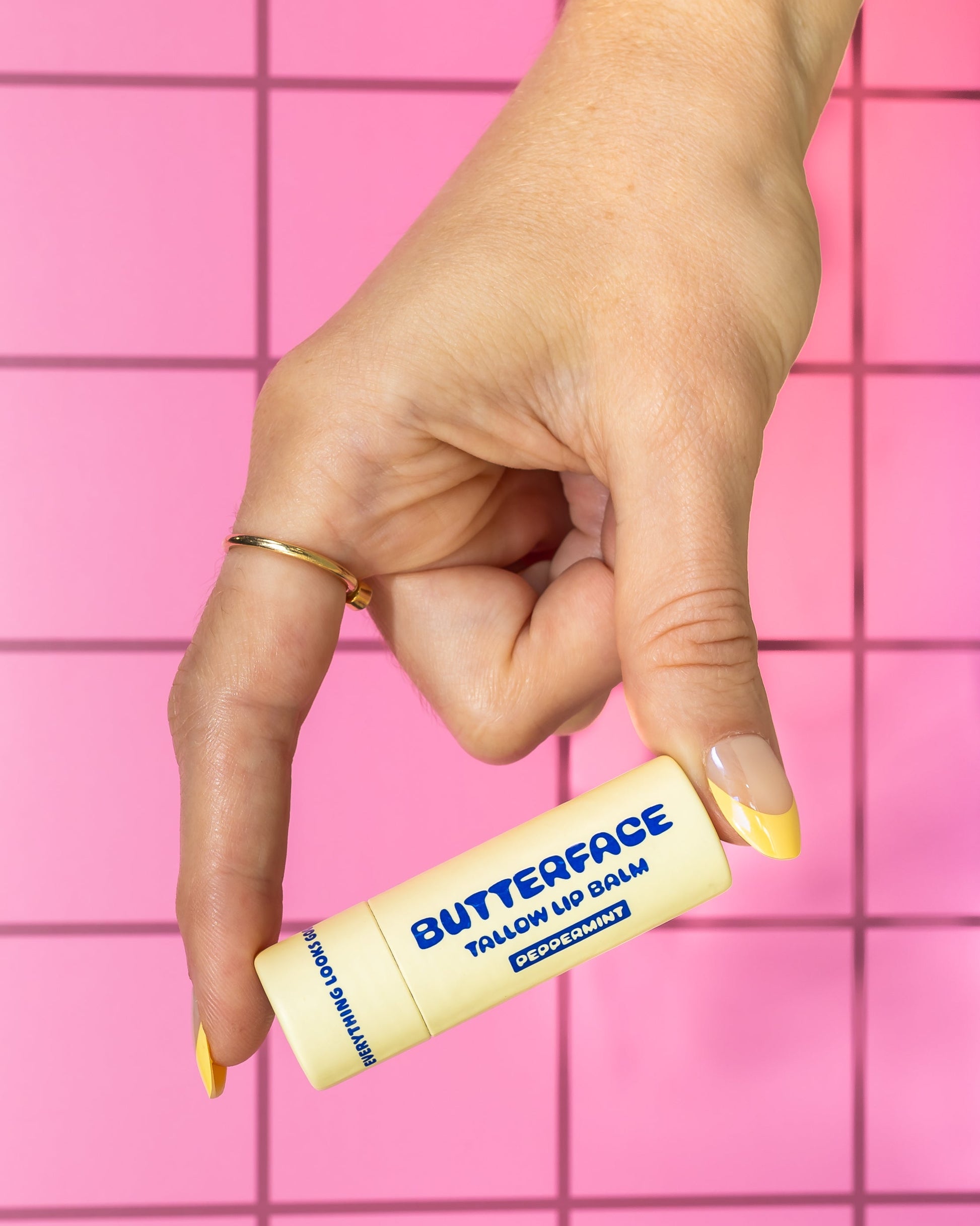 Hand holding a tallow butter lip balm named Butterface in a plastic-free tube against a pink tiled background